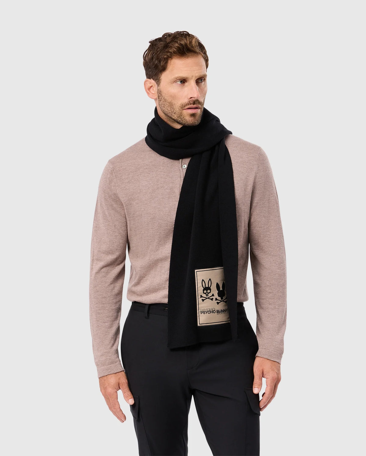 A man with short brown hair and a beard is wearing a beige long-sleeve shirt and black pants. He has a Psycho Bunny MENS MONROE TWILL PATCH SCARF - B6A892D200 in black draped around his neck, featuring a beige patch displaying two black outlined animated characters. He is standing against a light gray background.