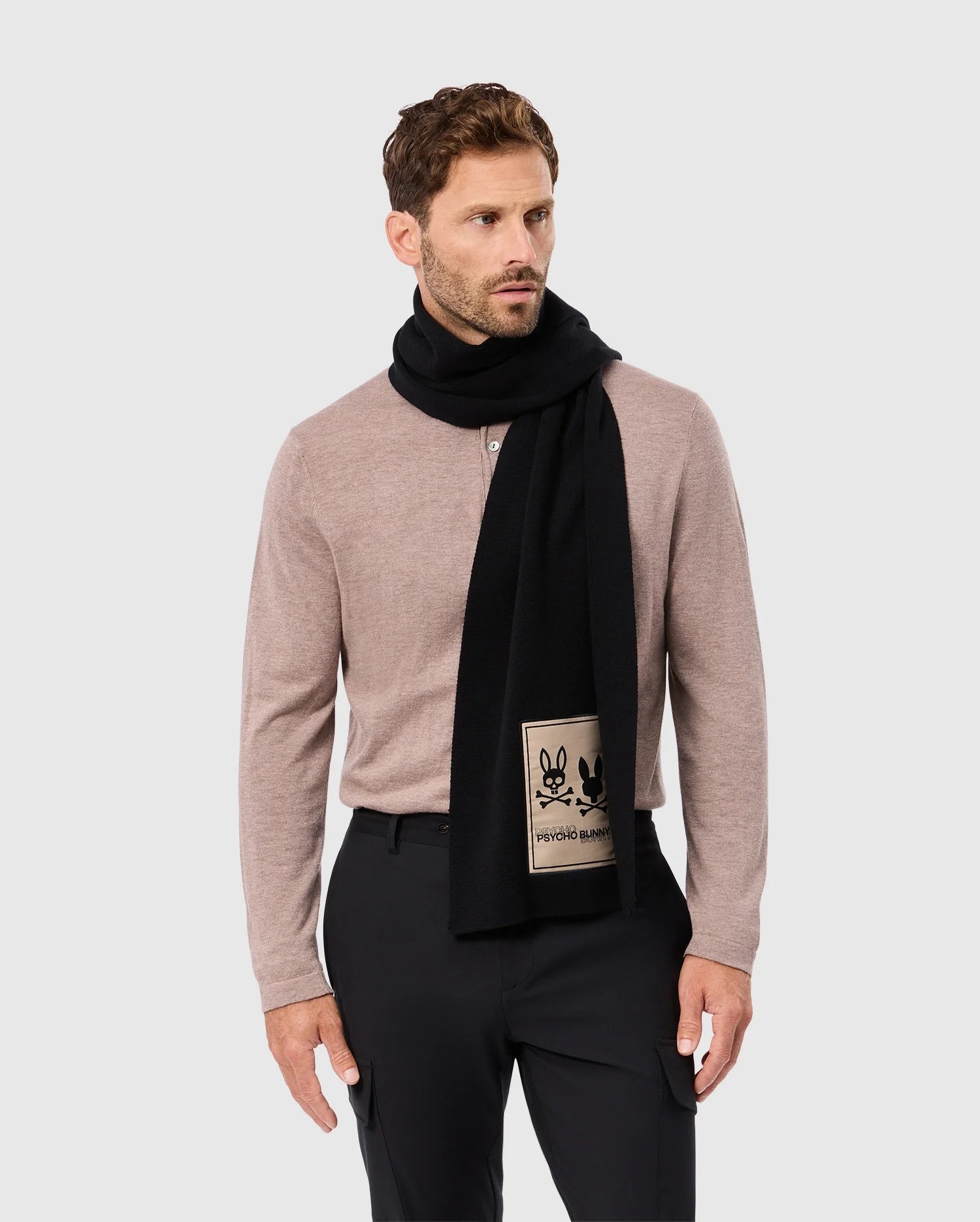 A man with short brown hair and a beard is wearing a beige long-sleeve shirt and black pants. He has a Psycho Bunny MENS MONROE TWILL PATCH SCARF - B6A892D200 in black draped around his neck, featuring a beige patch displaying two black outlined animated characters. He is standing against a light gray background.