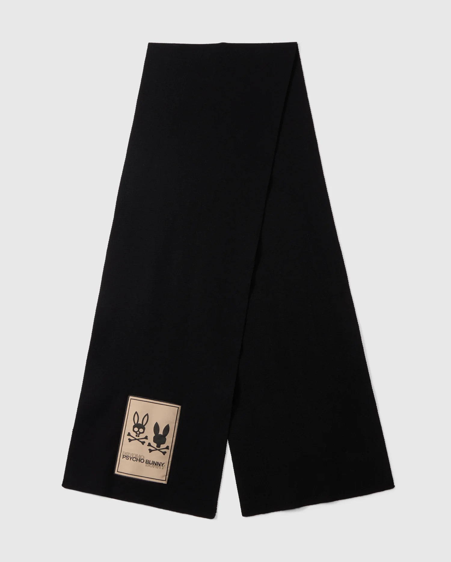 A long men's black MENS MONROE TWILL PATCH SCARF - B6A892D200 by Psycho Bunny is laid flat against a white background. This luxury scarf features a rectangular beige patch near one end, displaying two outlined bunny faces along with the words 