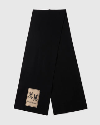 A long men's black MENS MONROE TWILL PATCH SCARF - B6A892D200 by Psycho Bunny is laid flat against a white background. This luxury scarf features a rectangular beige patch near one end, displaying two outlined bunny faces along with the words "PSYCHO BUNNY" at the bottom of the patch.