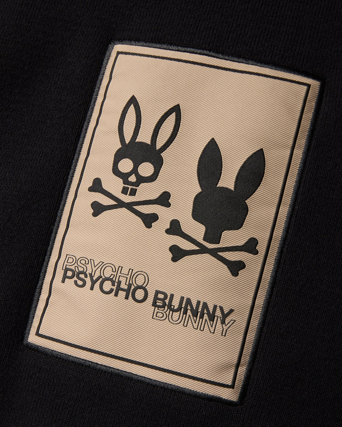 The MENS MONROE TWILL PATCH SCARF - B6A892D200 by Psycho Bunny features a black rectangular patch that displays two bunny skull logos with crossbones underneath. The left bunny skull has a cheerful, open-mouthed expression while the right one has a neutral look. Below the logos, "PSYCHO BUNNY" is written twice in uppercase letters.