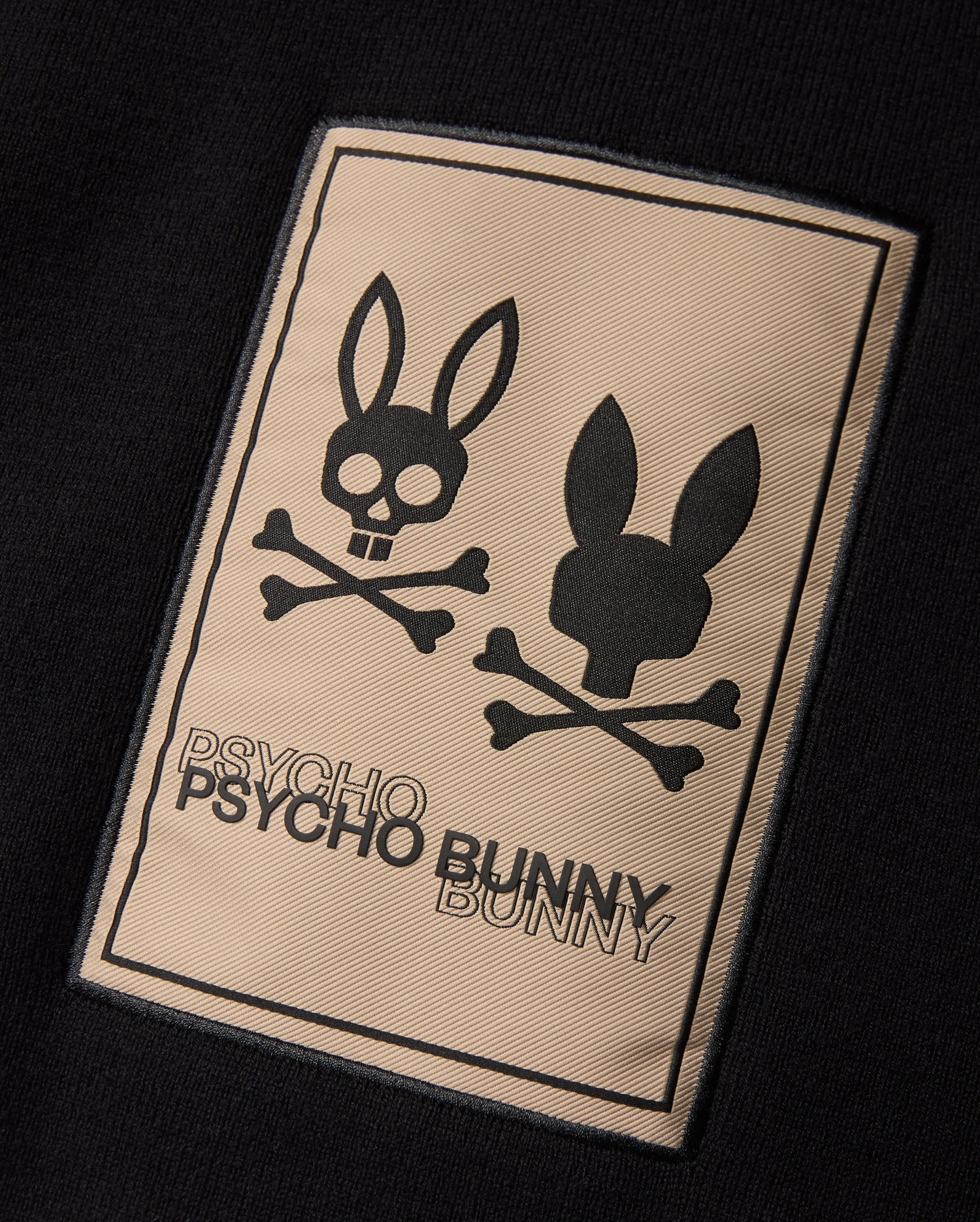 The MENS MONROE TWILL PATCH SCARF - B6A892D200 by Psycho Bunny features a black rectangular patch that displays two bunny skull logos with crossbones underneath. The left bunny skull has a cheerful, open-mouthed expression while the right one has a neutral look. Below the logos, "PSYCHO BUNNY" is written twice in uppercase letters.
