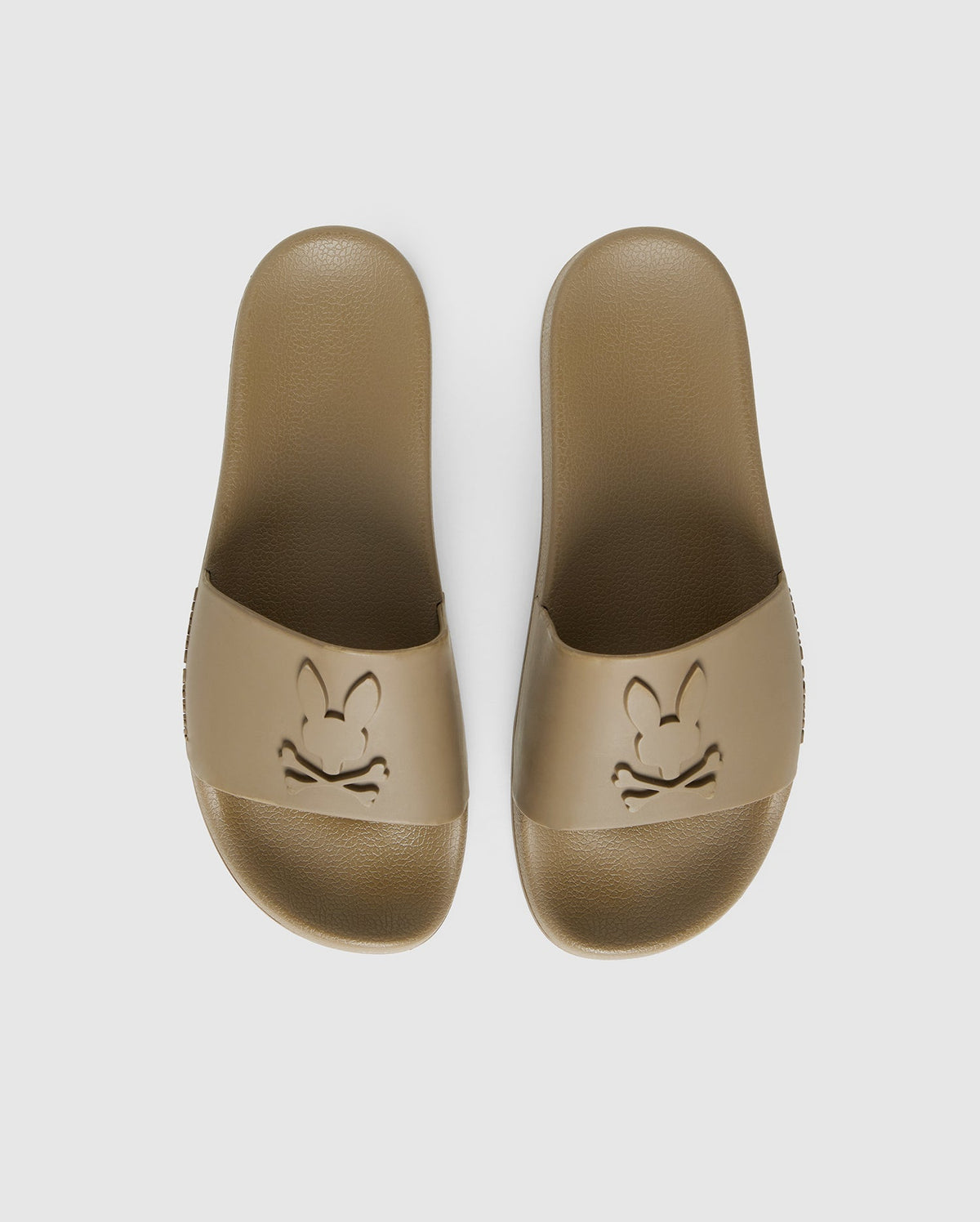 A pair of beige pool slides with a textured sole and upper strap featuring an embossed design. The design consists of a stylized bunny face with crossbones underneath, creating a playful skull and crossbones motif. They come with a molded footbed for added comfort and support, against a plain white background. 

Product Name: MENS LOGO SLIDES - B6A910U1FT 
Brand Name: Psycho Bunny