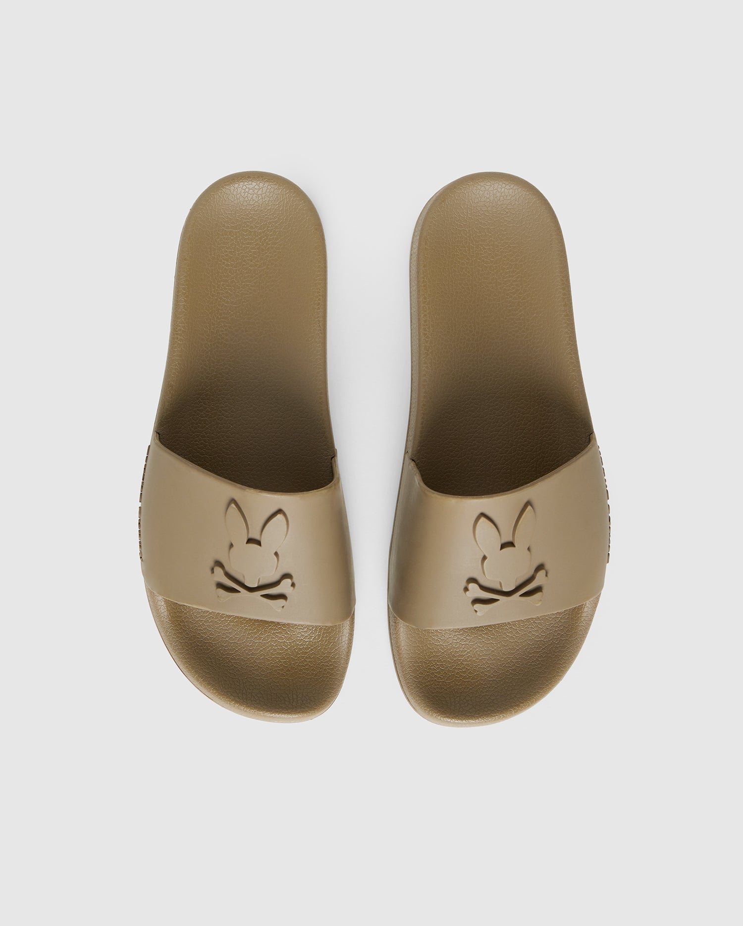 A pair of beige pool slides with a textured sole and upper strap featuring an embossed design. The design consists of a stylized bunny face with crossbones underneath, creating a playful skull and crossbones motif. They come with a molded footbed for added comfort and support, against a plain white background. 

Product Name: MENS LOGO SLIDES - B6A910U1FT 
Brand Name: Psycho Bunny