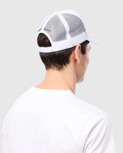 A person is seen from behind wearing a Psycho Bunny MENS BUNNY PATCH TRUCKER CAP - B6A965C200 with breathable mesh and a Bunny logo patch, paired with a white T-shirt, against a solid light gray background.