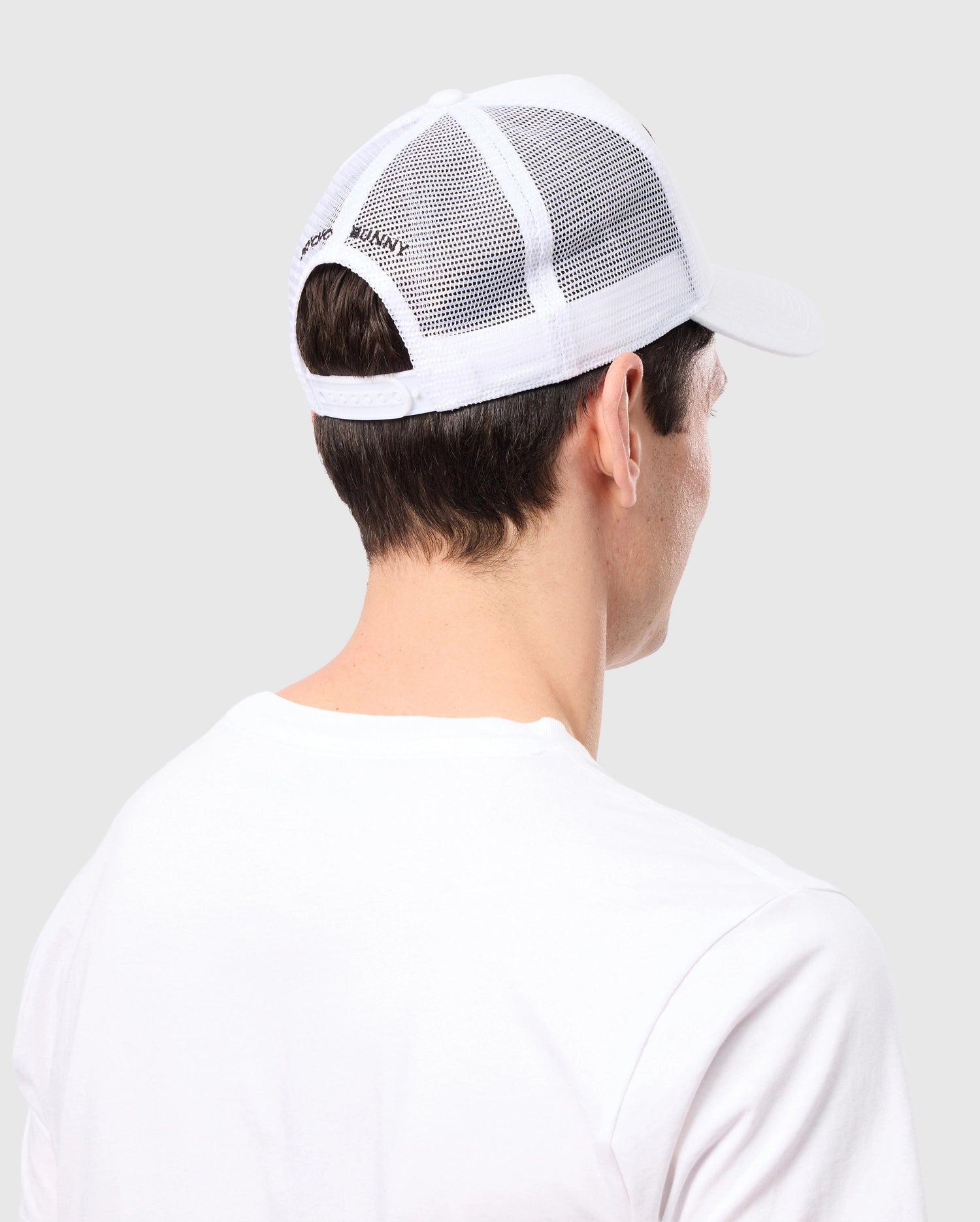 A person is seen from behind wearing a Psycho Bunny MENS BUNNY PATCH TRUCKER CAP - B6A965C200 with breathable mesh and a Bunny logo patch, paired with a white T-shirt, against a solid light gray background.