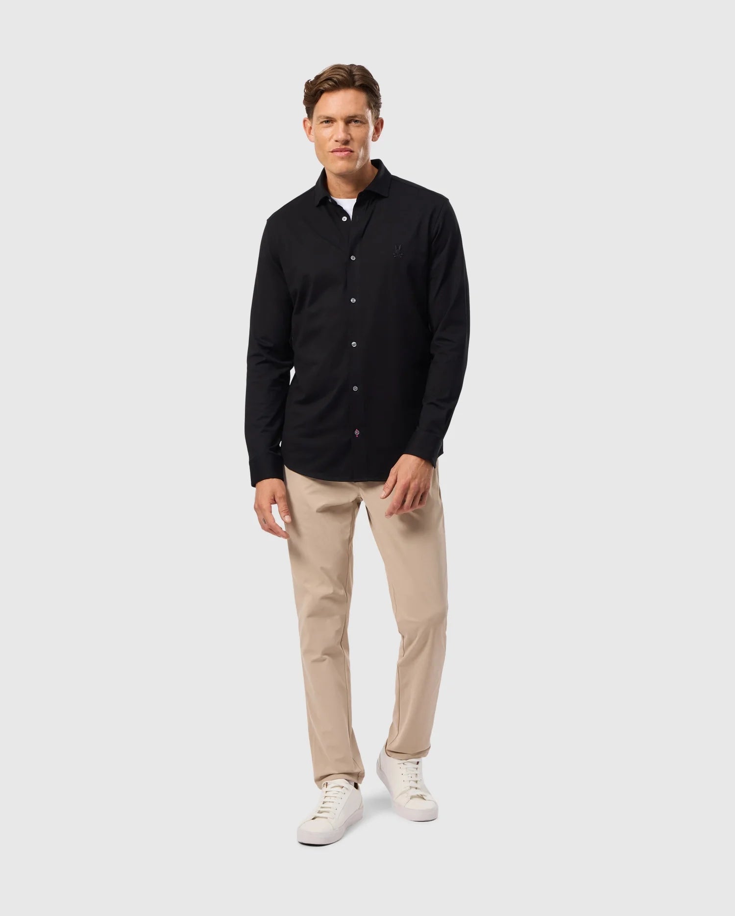 A person is standing against a plain background, wearing a Psycho Bunny MENS ARTHUR JERSEY SHIRT - B6C748D200 made of stretch Pima cotton, beige trousers, and white sneakers. They have short hair and are smiling slightly, with one hand hanging by their side and the other partially in a pocket.