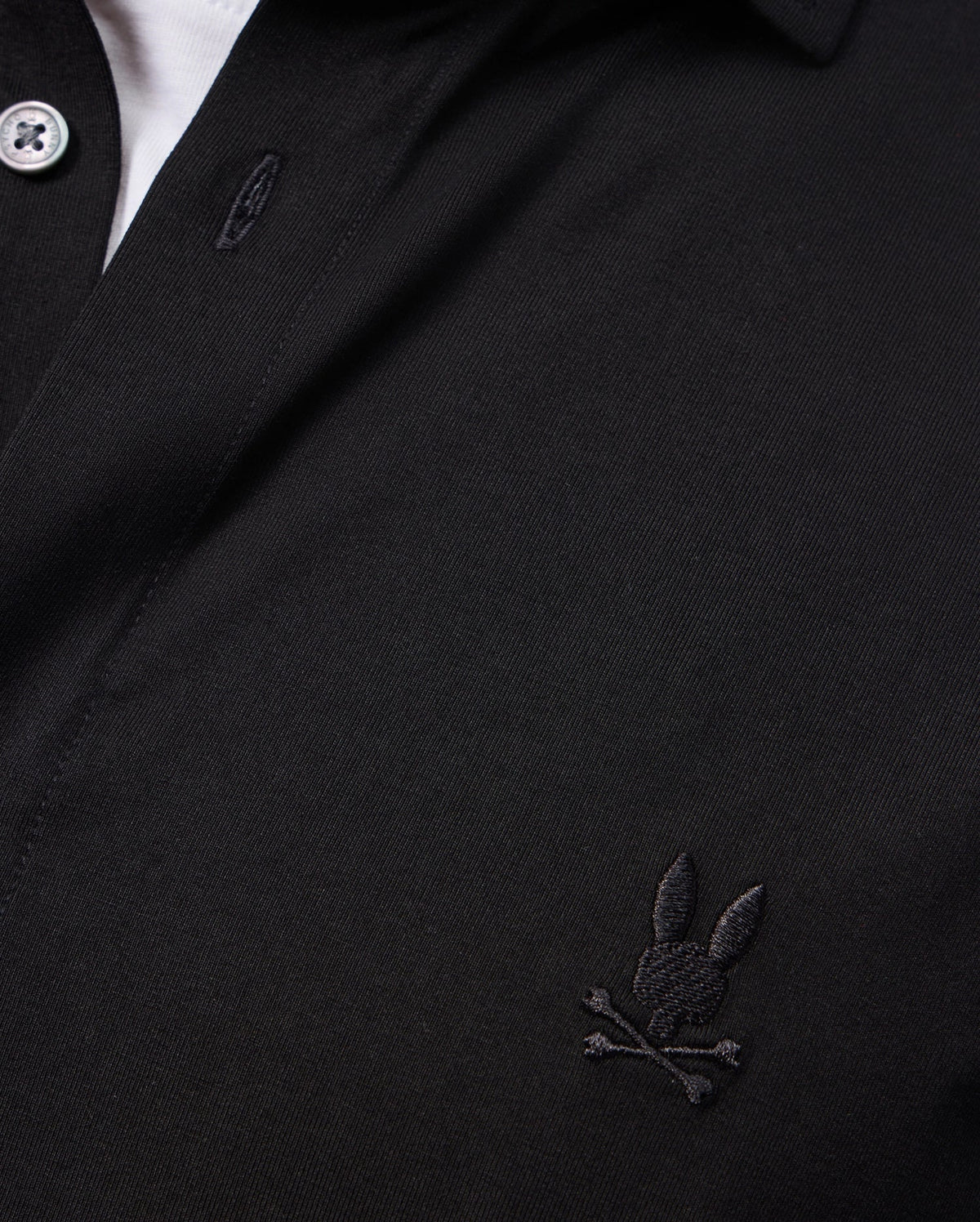 Close-up of the Psycho Bunny MENS ARTHUR JERSEY SHIRT - B6C748D200 in rich black, showcasing a small embroidered logo on the chest. Made from stretch Pima cotton jersey fabric, the playful design features a rabbit's head with crossed bones, adding a unique twist to this classic look.