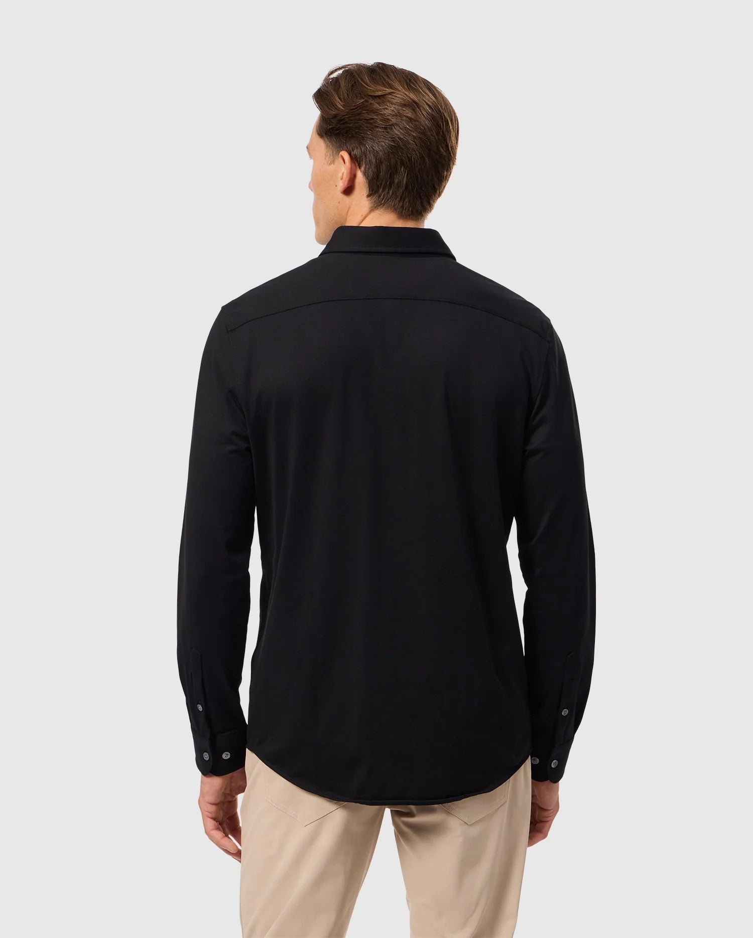 A man with short brown hair is shown from the back, wearing a black long-sleeved Psycho Bunny MENS ARTHUR JERSEY SHIRT - B6C748D200 and beige pants. The background is a plain light gray.