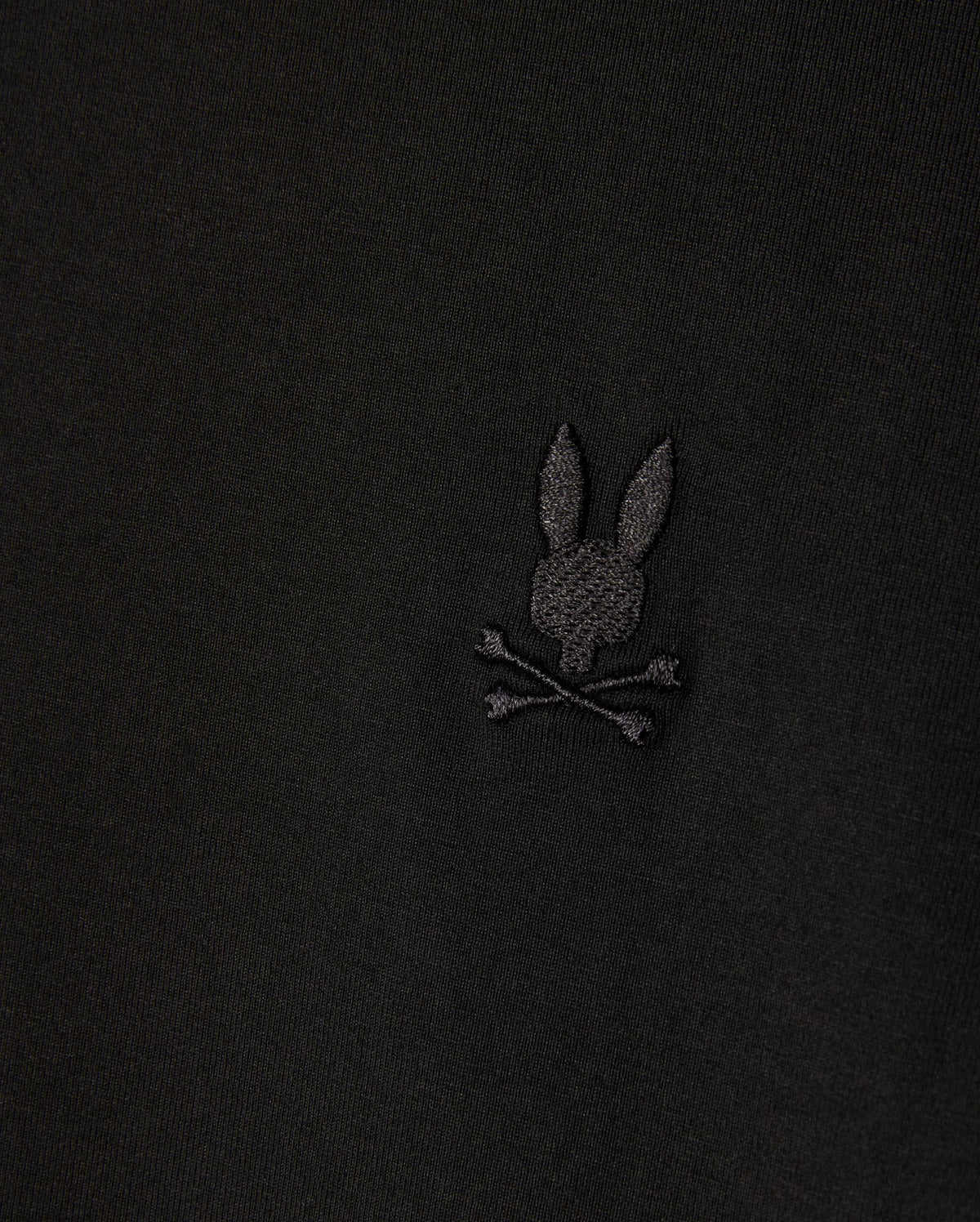Close-up of a Psycho Bunny MENS ARTHUR JERSEY SHIRT in stretch Pima cotton, displaying an embroidered bunny head with long ears above crossed bones, all set against black jersey fabric. (Product Code: B6C748D200).