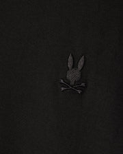 Close-up of a Psycho Bunny MENS ARTHUR JERSEY SHIRT in stretch Pima cotton, displaying an embroidered bunny head with long ears above crossed bones, all set against black jersey fabric. (Product Code: B6C748D200).