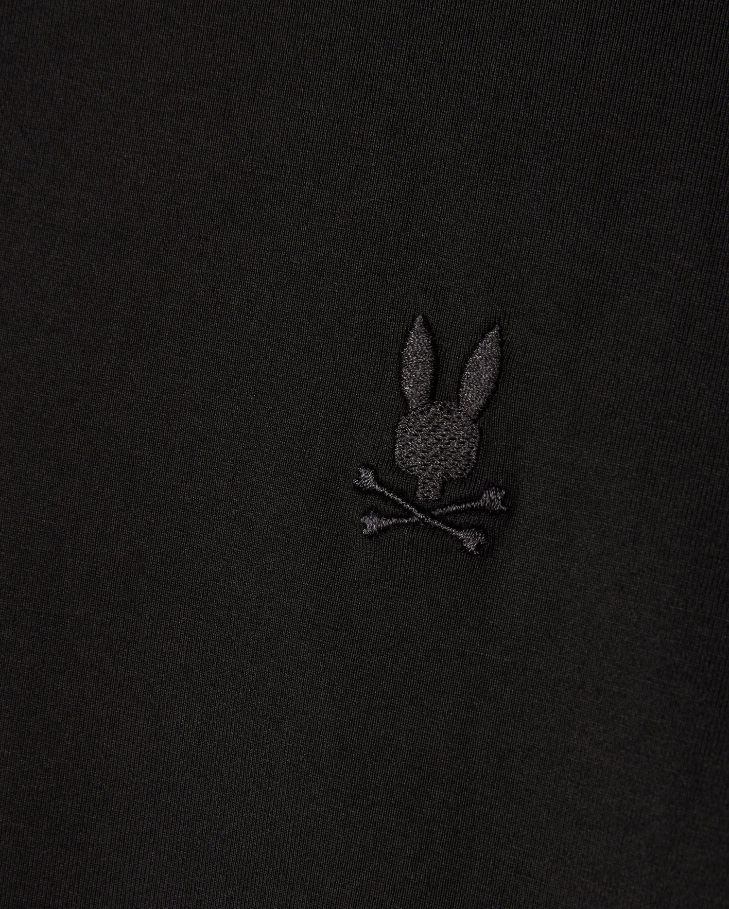 Close-up of a Psycho Bunny MENS ARTHUR JERSEY SHIRT in stretch Pima cotton, displaying an embroidered bunny head with long ears above crossed bones, all set against black jersey fabric. (Product Code: B6C748D200).