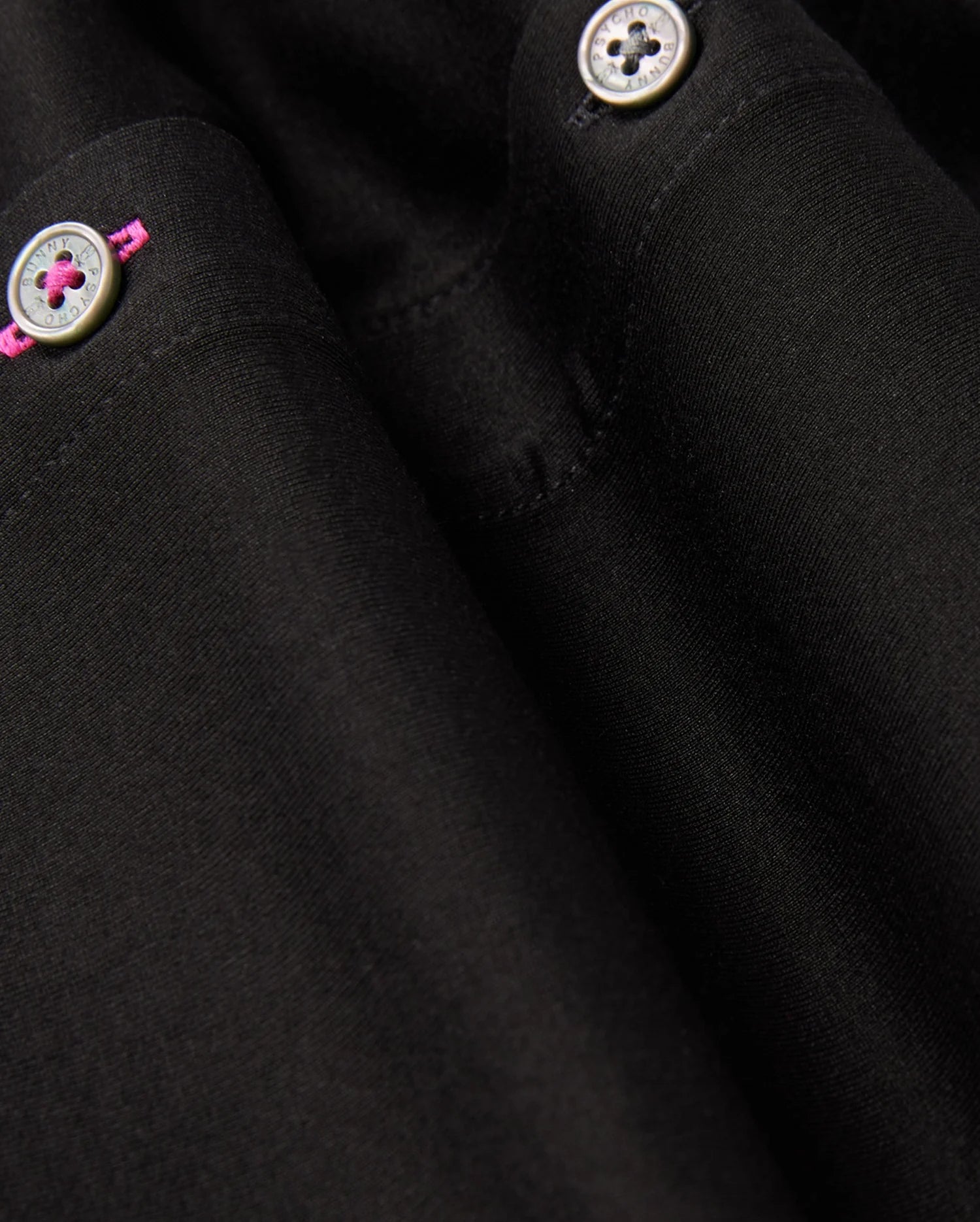 Close-up of the black fabric from Psycho Bunny's MENS ARTHUR JERSEY SHIRT - B6C748D200, featuring two small metallic buttons. The upper buttonhole is accentuated with a pink thread stitch, providing a subtle pop of color against the smooth, finely woven monochromatic textile.