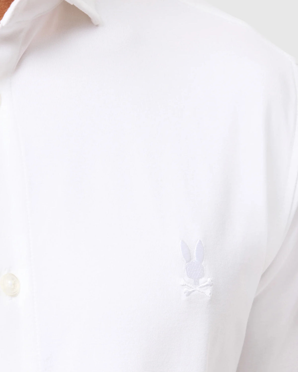 A close-up of the upper part of a MENS ARTHUR JERSEY SHIRT - B6C748D200 from Psycho Bunny, crafted in stretch Pima cotton, featuring a small embroidered logo on the left side. The logo consists of an outline of a bunny's head with crossed bones beneath it. A portion of the open collar and top button is also visible.