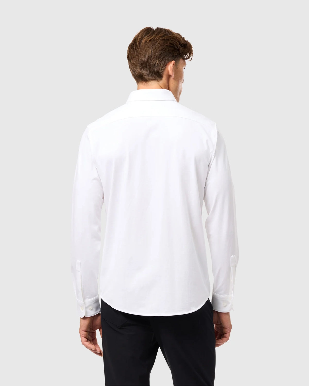 A person with short brown hair is standing with their back to the camera, wearing a Psycho Bunny Mens Arthur Jersey Shirt (B6C748D200) made from stretch Pima cotton, along with black pants. The background is a plain, light gray color.