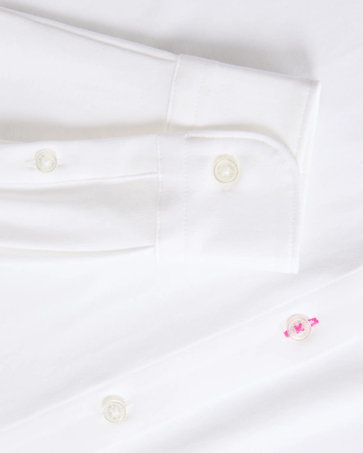 Close-up of a Psycho Bunny's Mens Arthur Jersey Shirt (B6C748D200) showcasing its buttoned cuff and two front buttons. Crafted from crisp, clean stretch Pima cotton, the fabric offers a subtle pop with one buttonhole stitched in pink thread, enhancing the elegance of this all-white shirt.