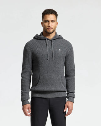 A man wears a Psycho Bunny MENS LEVAN WAFFLE HOODED SWEATER in gray, featuring an embroidered chest bunny, drawstring, and front pocket. He pairs it with black pants for a minimalistic look.
