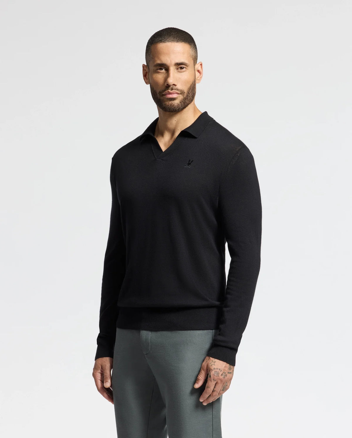 A bearded man wearing a Psycho Bunny MENS JOHNNY COLLAR SWEATER (B6E258E200) in black, paired with gray pants, stands against a light gray background.