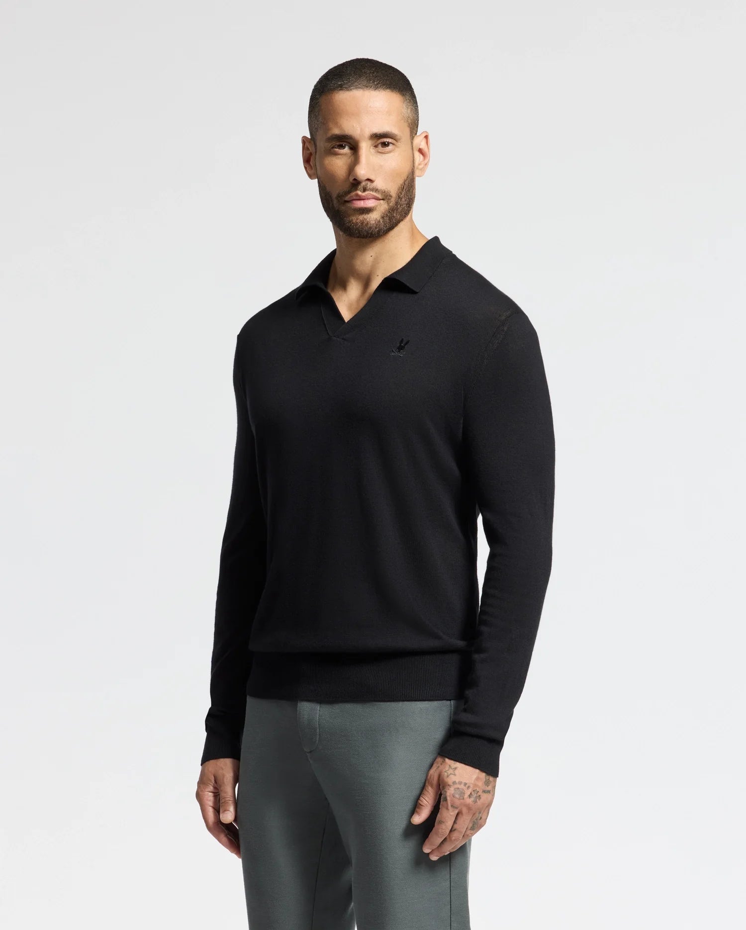 A bearded man wearing a Psycho Bunny MENS JOHNNY COLLAR SWEATER (B6E258E200) in black, paired with gray pants, stands against a light gray background.
