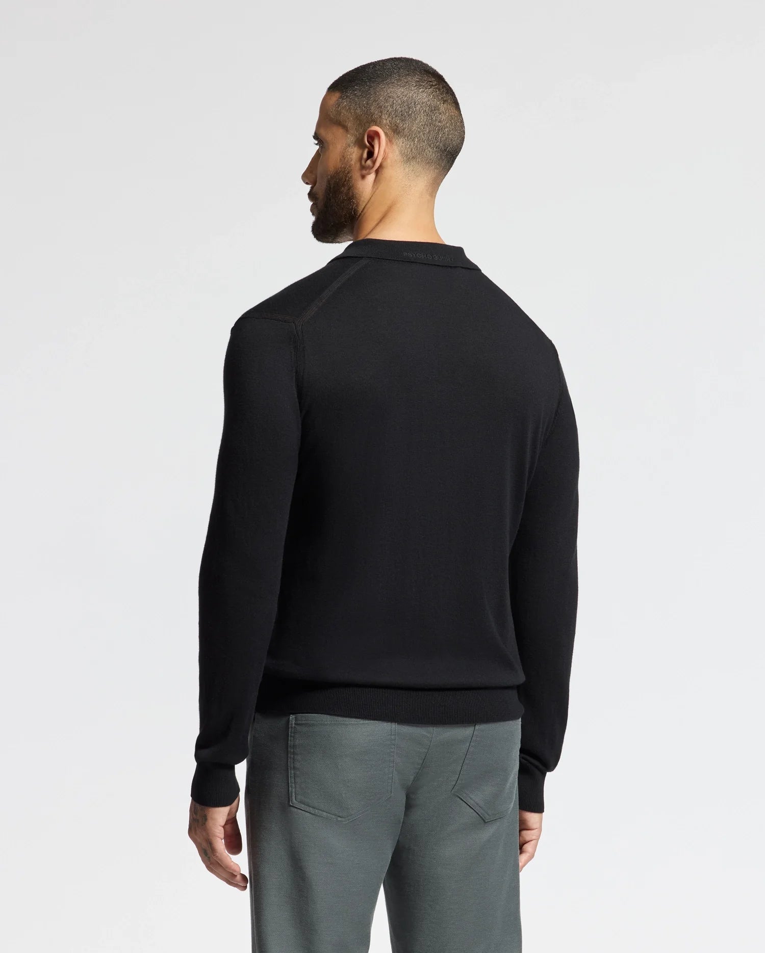 A man with short hair, slightly turned from the camera, wears a black Psycho Bunny MENS JOHNNY COLLAR SWEATER - B6E258E200 with gray pants. The simple cotton-wool blend attire contrasts elegantly against the plain light gray background.