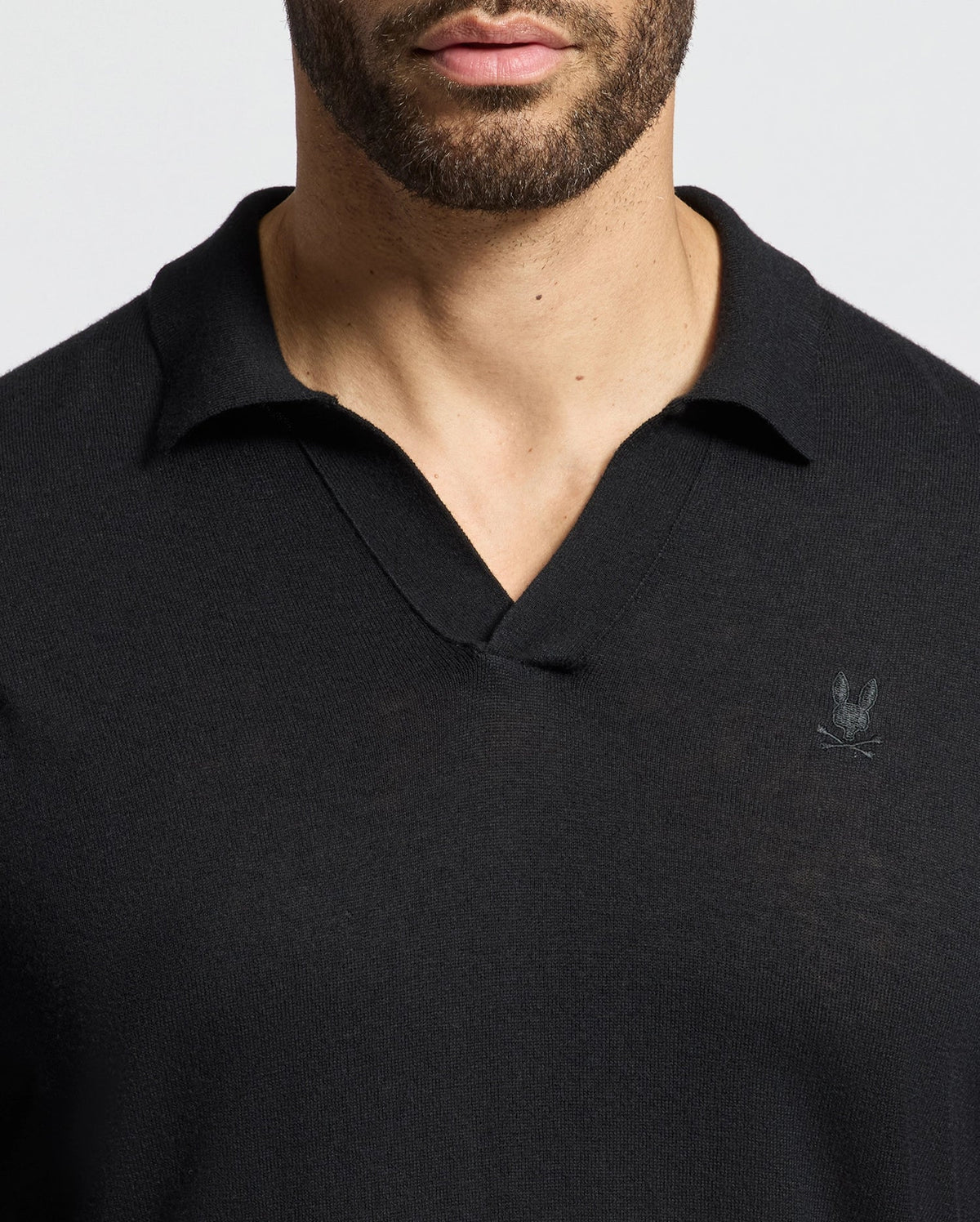 A person is wearing a Psycho Bunny men's Johnny Collar Sweater (B6E258E200), featuring a small embroidered rabbit head logo on the left chest. The cozy black V-neck is made from a cotton-wool blend, perfectly complementing the plain, light-colored background.