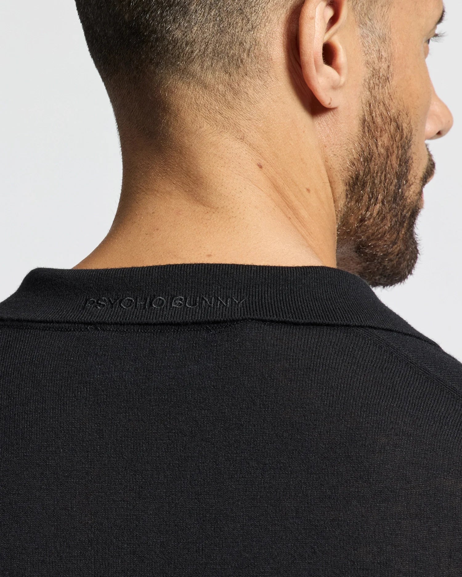 A man with short hair and a beard wears the black Psycho Bunny MENS JOHNNY COLLAR SWEATER, B6E258E200, featuring a subtle embossing near the neckline. This cotton-wool blend piece adds sophistication against a plain, light backdrop.