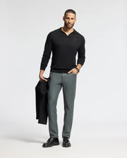 A man stands against a light background, wearing the Psycho Bunny MENS JOHNNY COLLAR SWEATER in black with gray trousers. He holds a black jacket in his right hand and pairs it with black shoes, exuding effortless style.