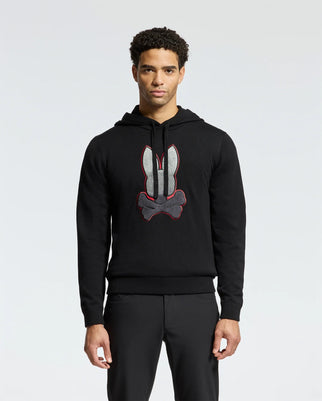 A man wearing Psycho Bunny's MENS BULDER HOODED SWEATER - B6E261E200, made from plush Supima cotton, stands against a plain background. The black hoodie showcases a bold bunny and crossbones graphic on the front.