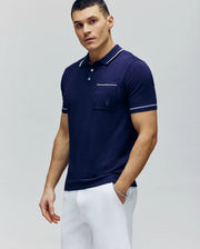 A man stands facing forward with one hand in his pocket, wearing Psycho Bunny’s MENS LONGFORD FINE KNIT POLO - B6E500F200 in navy blue with white trim and slim-fit white pants, set against a plain light background.