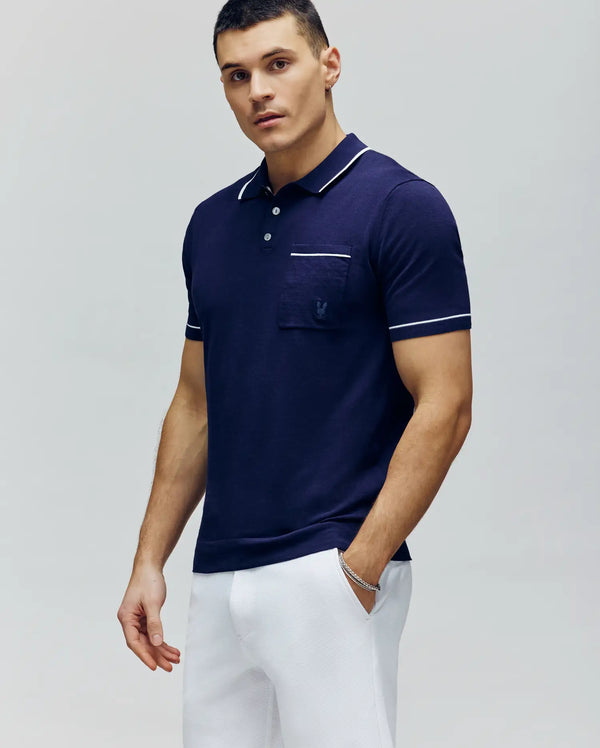 A man stands facing forward with one hand in his pocket, wearing Psycho Bunny’s MENS LONGFORD FINE KNIT POLO - B6E500F200 in navy blue with white trim and slim-fit white pants, set against a plain light background.