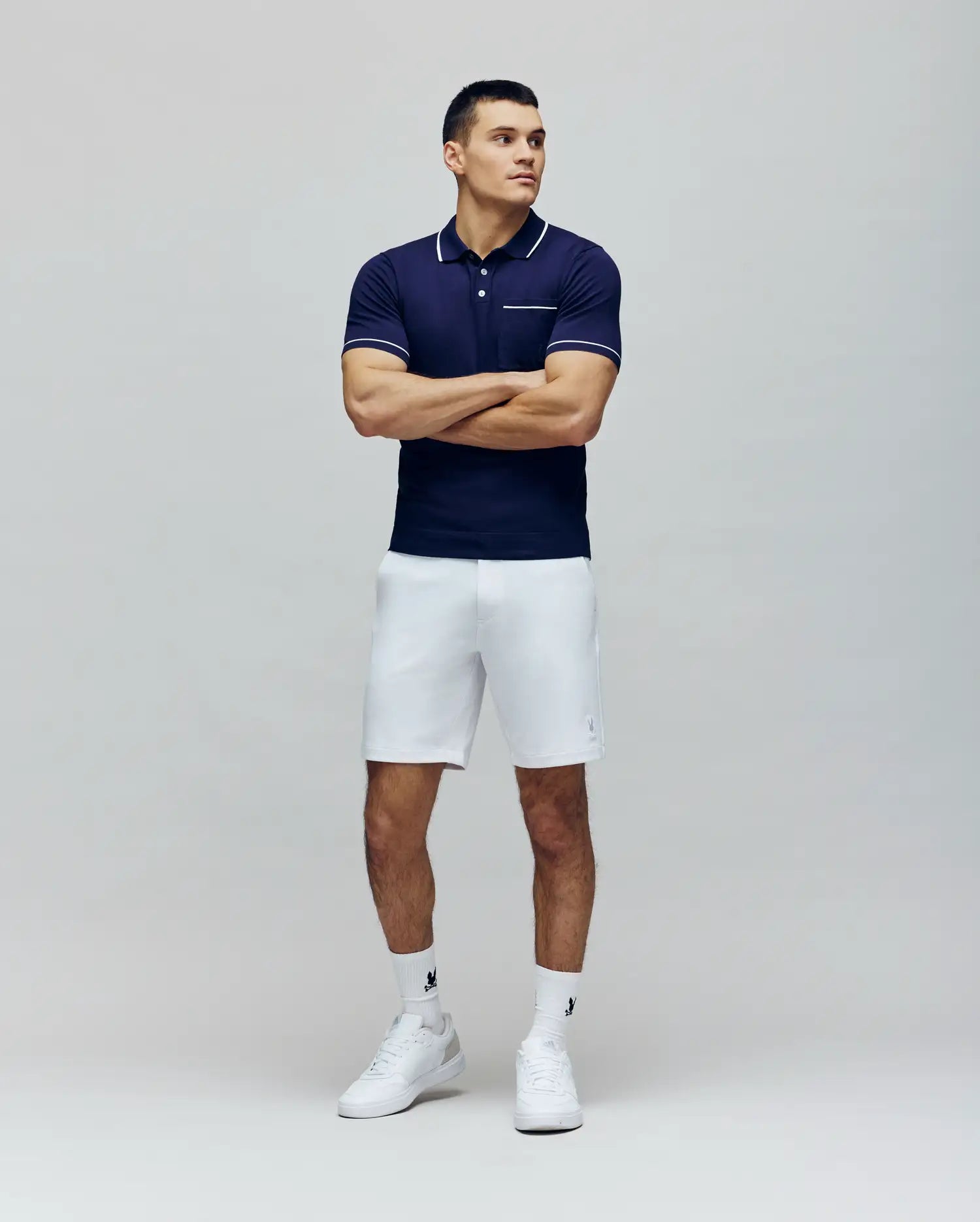 A man stands with arms crossed wearing the Psycho Bunny MENS LONGFORD FINE KNIT POLO - B6E500F200 in navy with white trim, paired with white shorts, crew socks, and sneakers against a light gray background.
