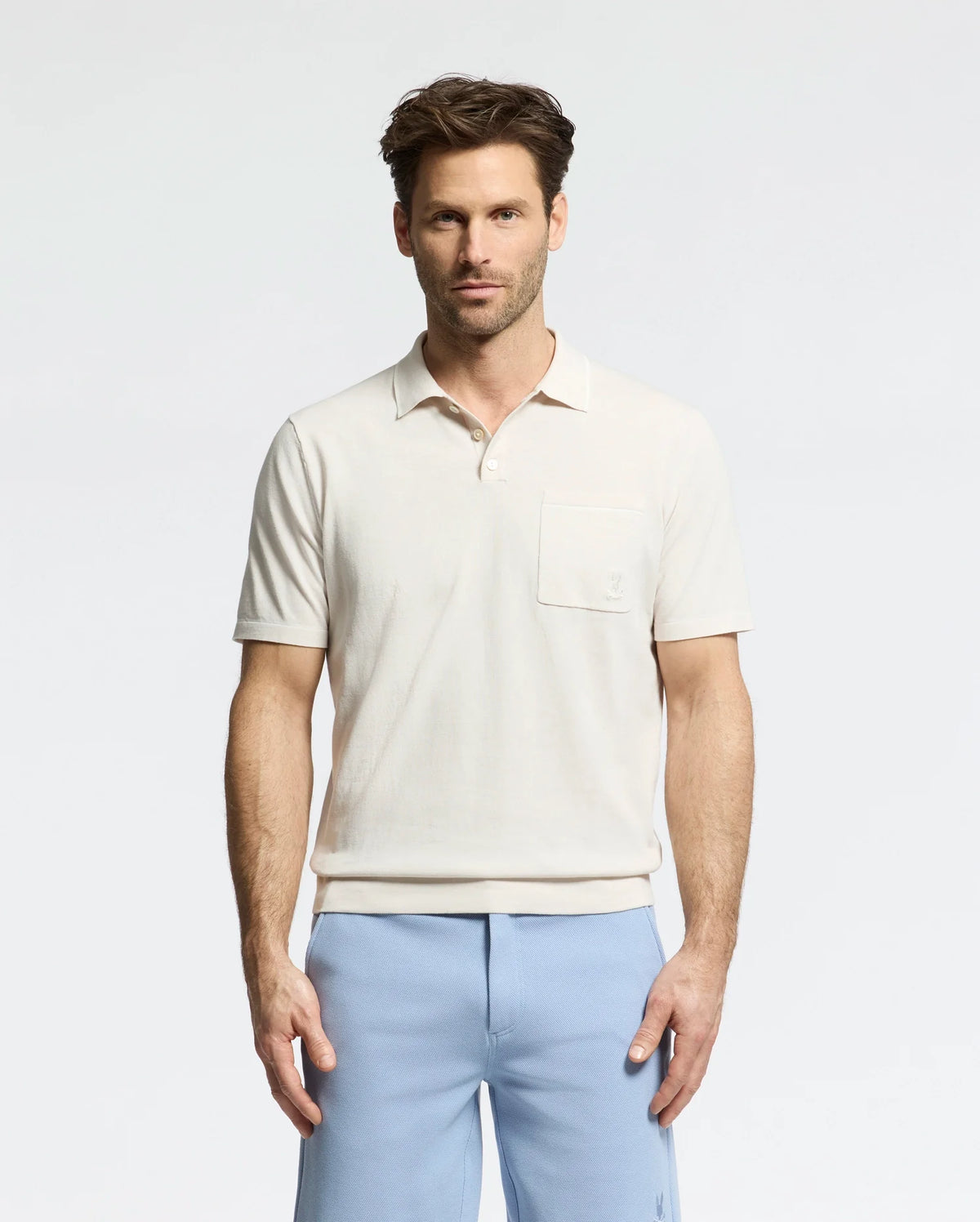 A man stands against a plain background wearing the Psycho Bunny MENS LONGFORD FINE KNIT POLO (B6E500F200) with light blue pants. With short, styled hair, he maintains a neutral expression while facing the camera.