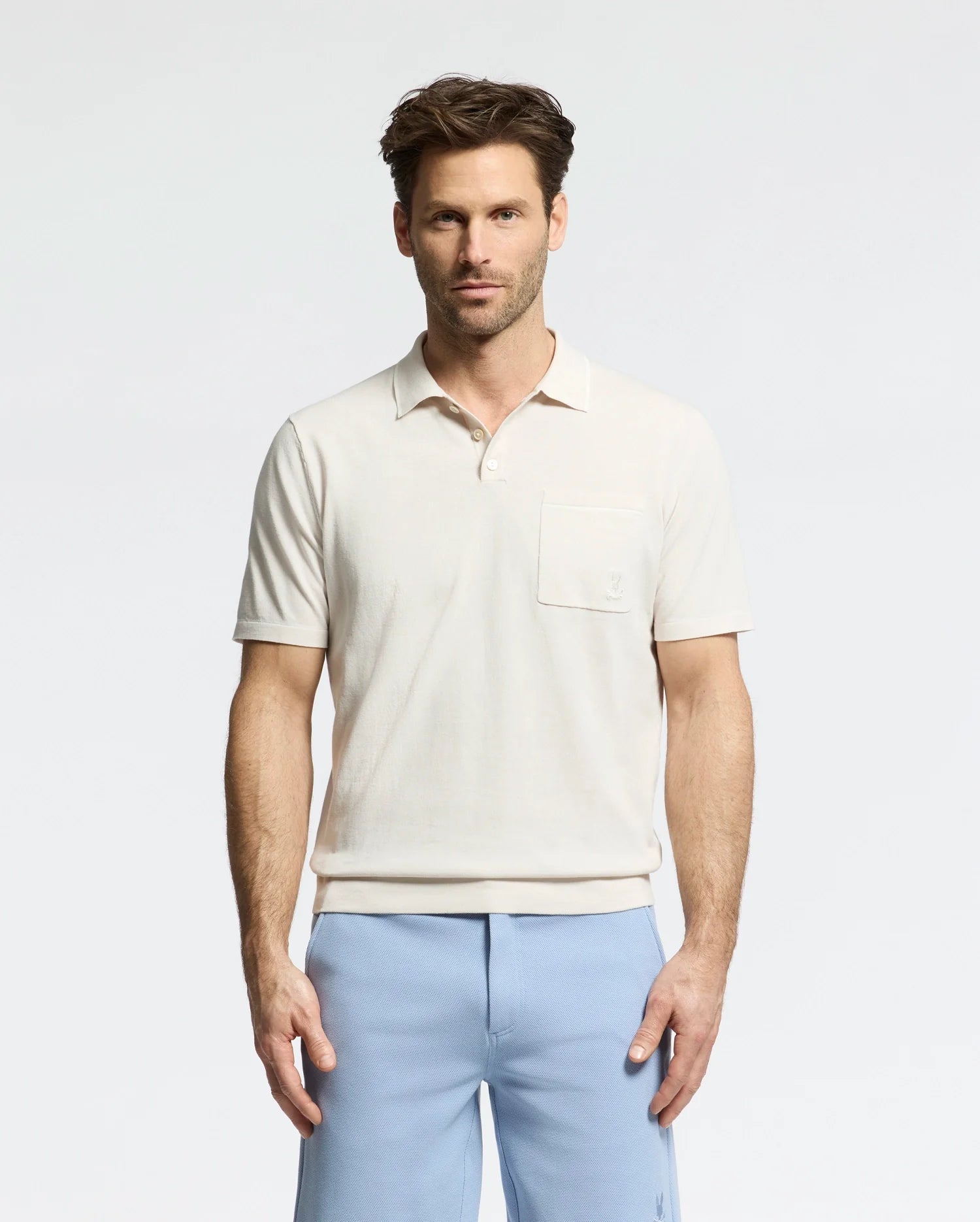 A man stands against a plain background wearing the Psycho Bunny MENS LONGFORD FINE KNIT POLO (B6E500F200) with light blue pants. With short, styled hair, he maintains a neutral expression while facing the camera.