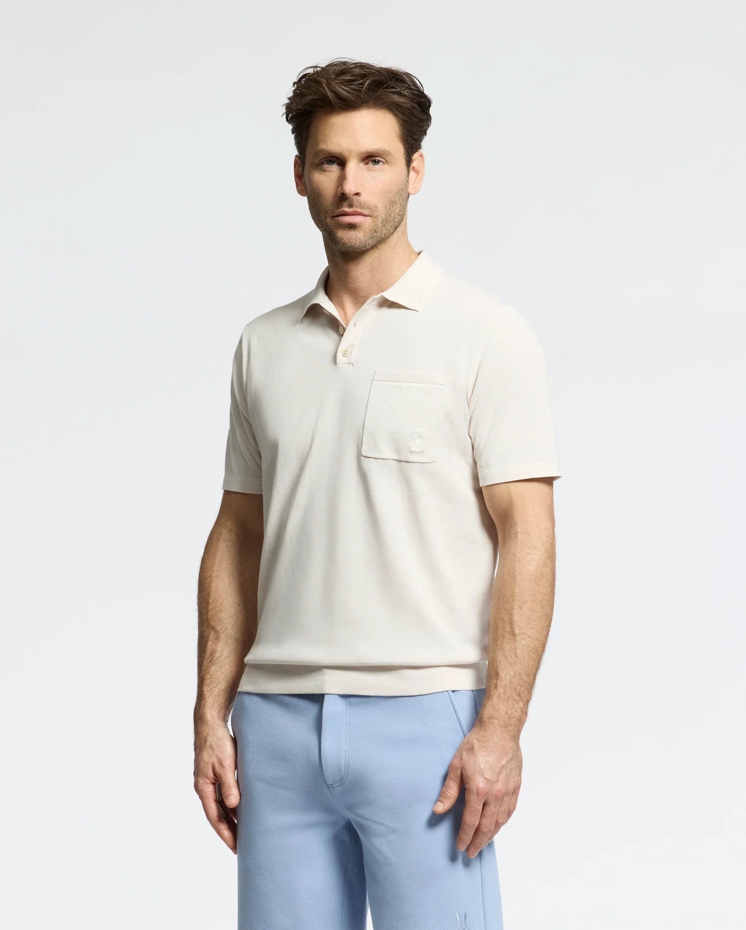 A man stands in a beige short-sleeved Psycho Bunny MENS LONGFORD FINE KNIT POLO - B6E500F200 with a chest pocket, paired with light blue pants against a plain white background.