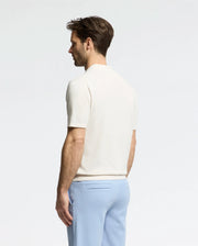 A man with short hair stands sideways wearing a Psycho Bunny MENS LONGFORD FINE KNIT POLO and light blue pants, set against a plain background that highlights his understated elegance.