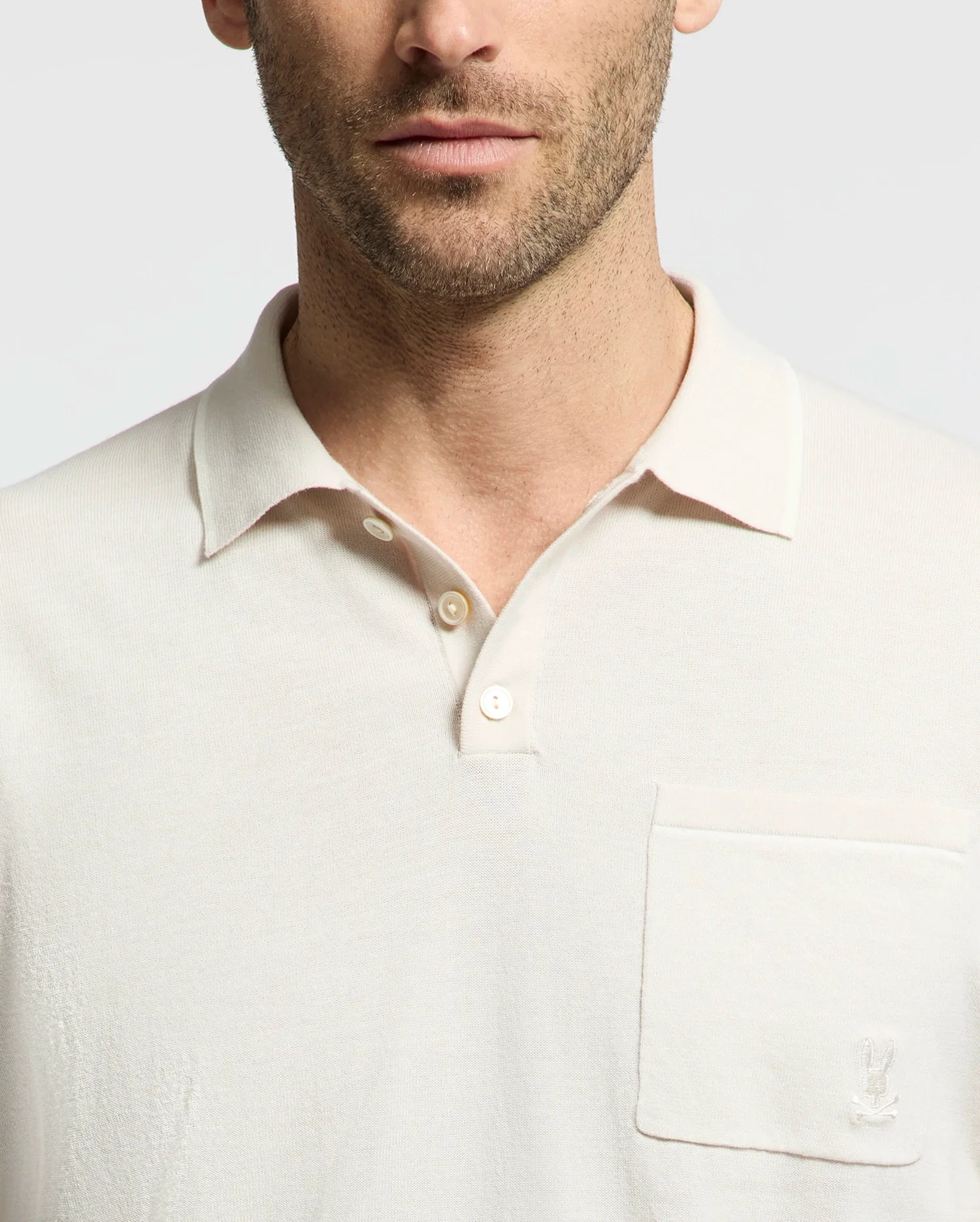A man wears a Psycho Bunny MENS LONGFORD FINE KNIT POLO in light cotton, featuring a collar and left chest pocket with subtle embroidered Bunny. The plain white design highlights his partially visible face.