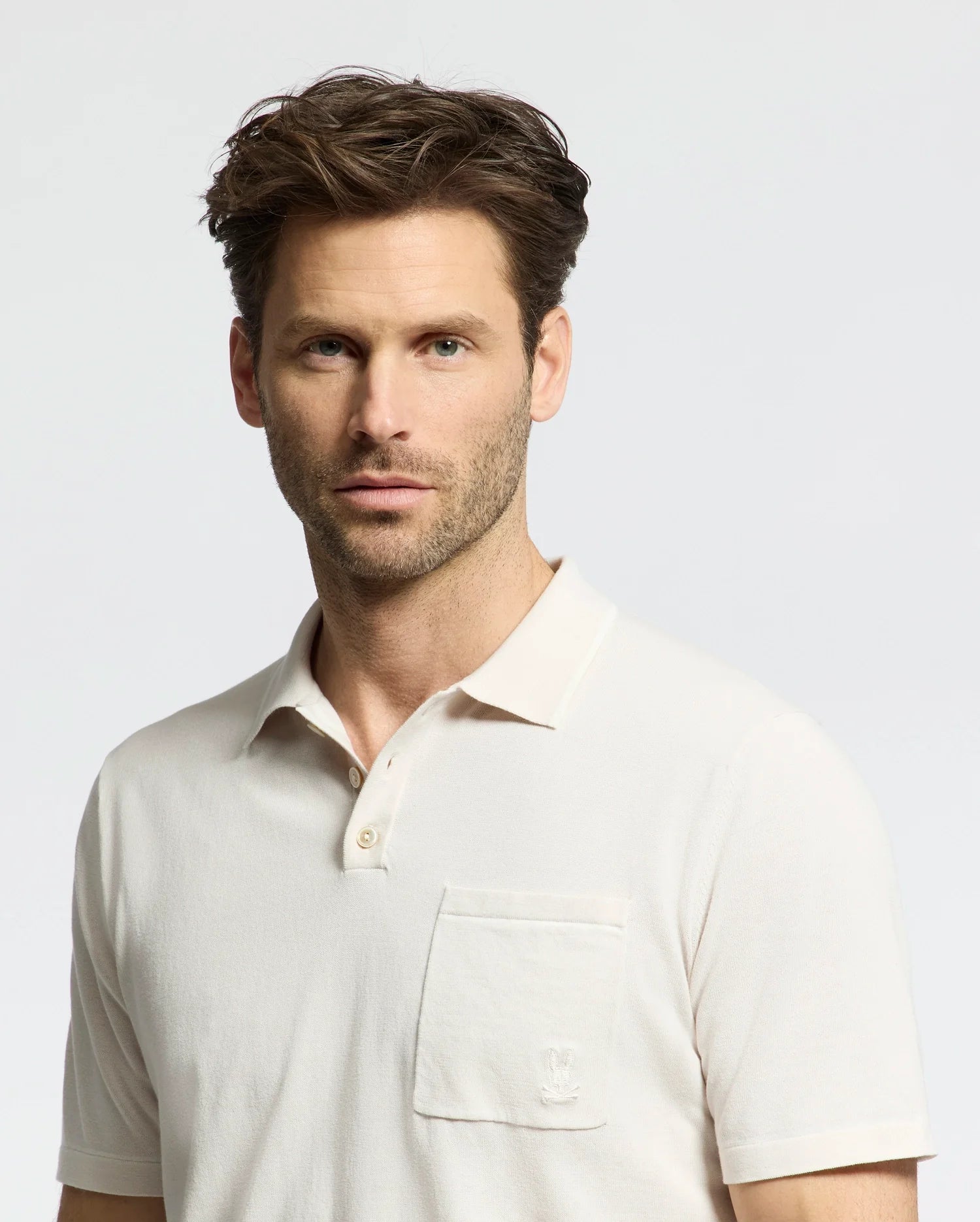 A man with short brown hair and light stubble wears a cream-colored Psycho Bunny MENS LONGFORD FINE KNIT POLO - B6E500F200, featuring an embroidered Bunny on the chest pocket. He stands against a plain light background.