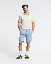 A man stands with hands in pockets wearing a Psycho Bunny MENS LONGFORD FINE KNIT POLO - B6E500F200 in light beige, paired with light blue shorts, white socks, and blue sneakers. The background is plain white, and he gazes slightly to the side.