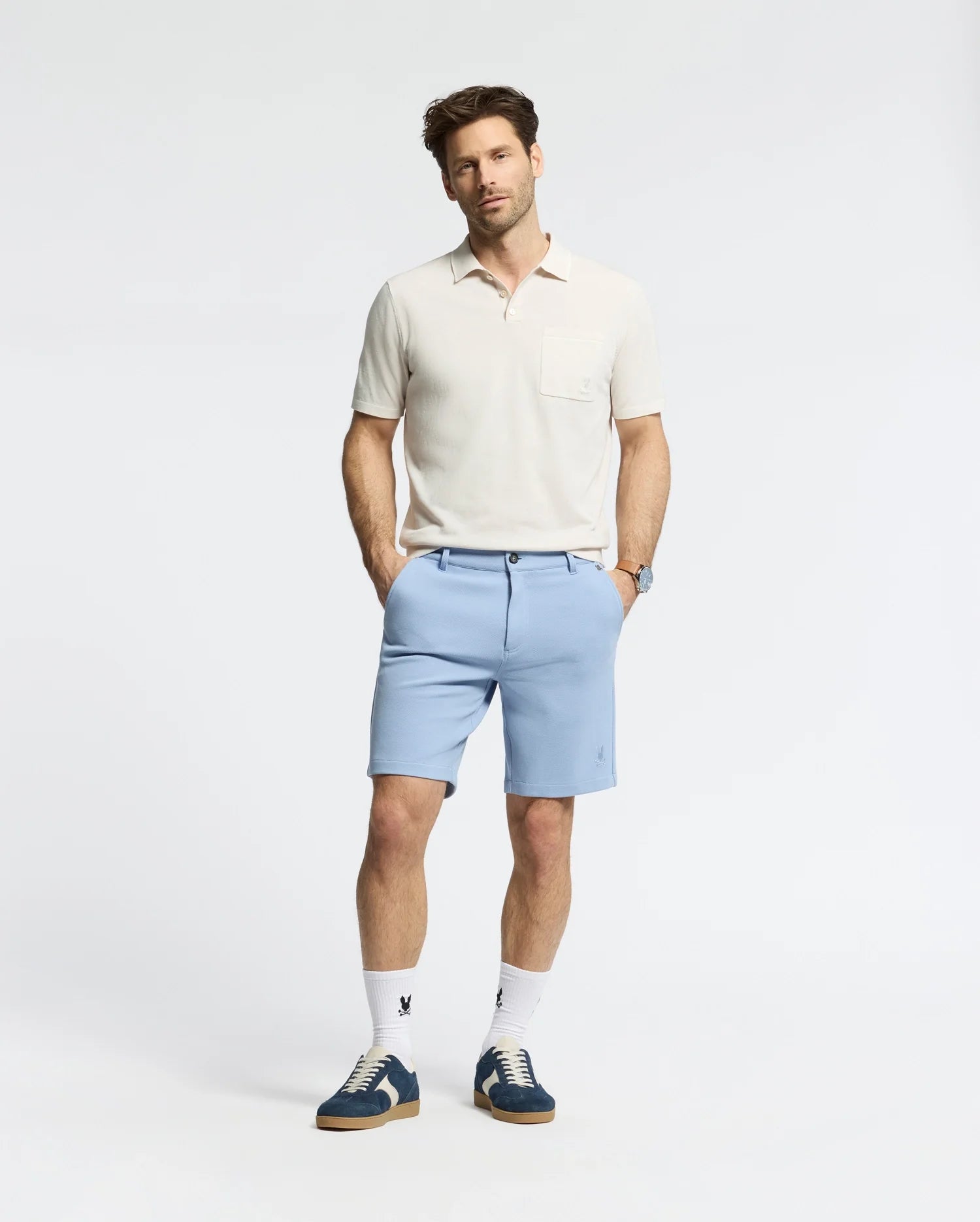 A man stands with hands in pockets wearing a Psycho Bunny MENS LONGFORD FINE KNIT POLO - B6E500F200 in light beige, paired with light blue shorts, white socks, and blue sneakers. The background is plain white, and he gazes slightly to the side.