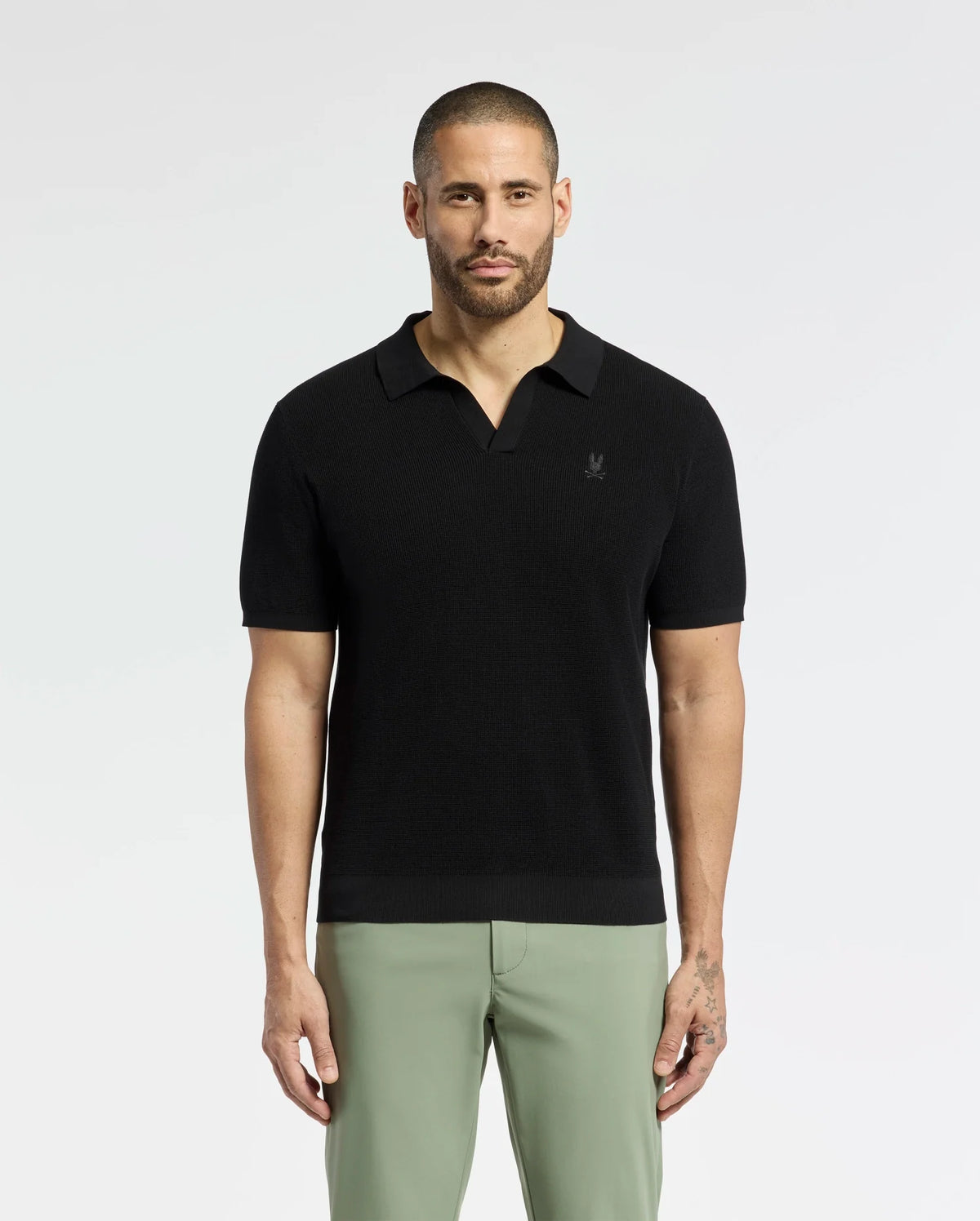 A bearded man confidently wears a breathable Psycho Bunny MENS LEETON JOHNNY COLLAR KNIT POLO (B6E562F200) in black, paired with light green pants. With an embroidered bunny, the 100% cotton shirt fits him perfectly as he gazes directly at the camera, showcasing his left-hand tattoo against a plain backdrop.
