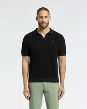 A bearded man confidently wears a breathable Psycho Bunny MENS LEETON JOHNNY COLLAR KNIT POLO (B6E562F200) in black, paired with light green pants. With an embroidered bunny, the 100% cotton shirt fits him perfectly as he gazes directly at the camera, showcasing his left-hand tattoo against a plain backdrop.