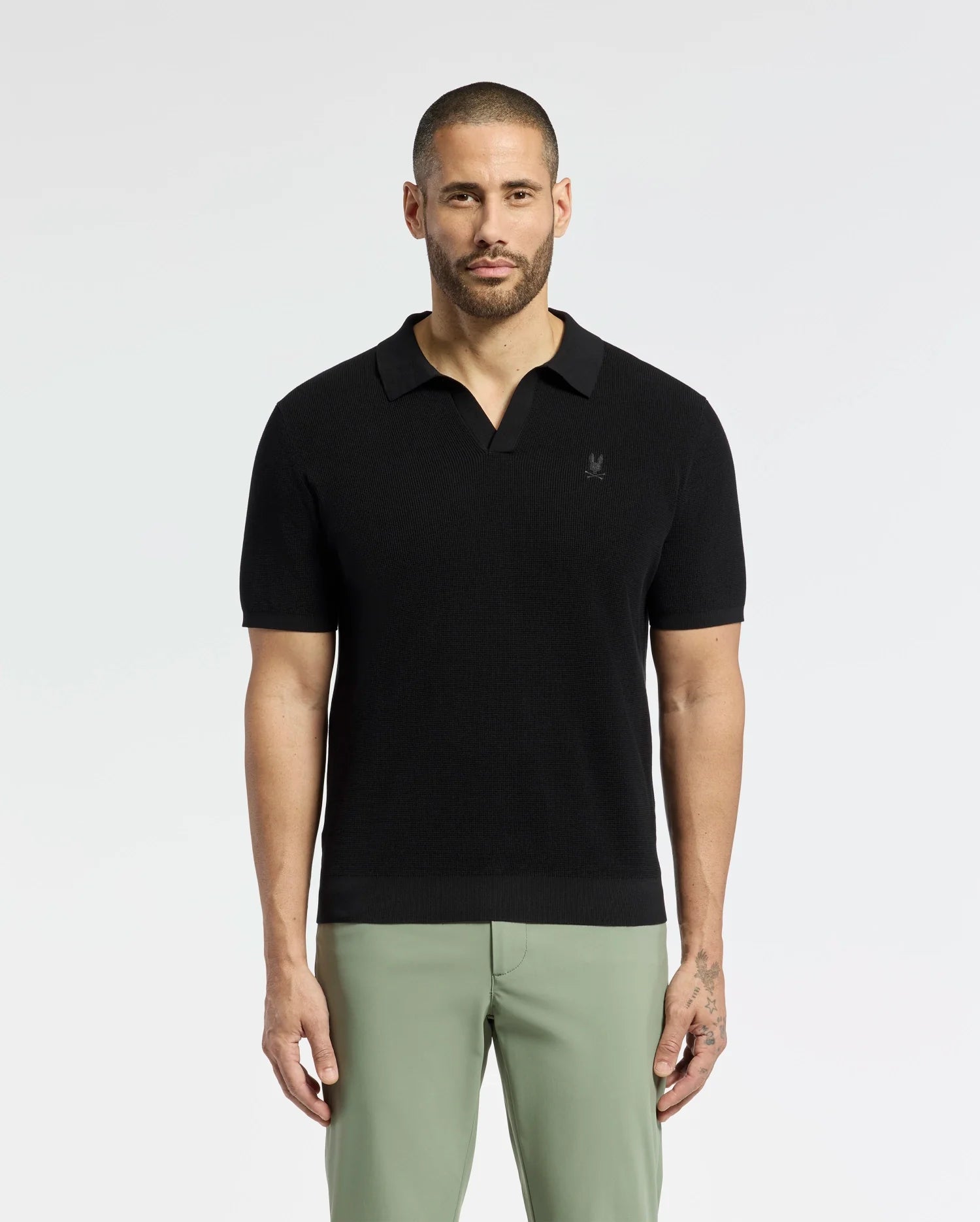 A bearded man confidently wears a breathable Psycho Bunny MENS LEETON JOHNNY COLLAR KNIT POLO (B6E562F200) in black, paired with light green pants. With an embroidered bunny, the 100% cotton shirt fits him perfectly as he gazes directly at the camera, showcasing his left-hand tattoo against a plain backdrop.