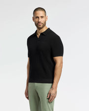 A bearded person stands against a plain backdrop in a black MENS LEETON JOHNNY COLLAR KNIT POLO by Psycho Bunny with light green pants. Their 100% breathable cotton polo features an embroidered Bunny, and tattoos are visible on one arm as they face the camera.