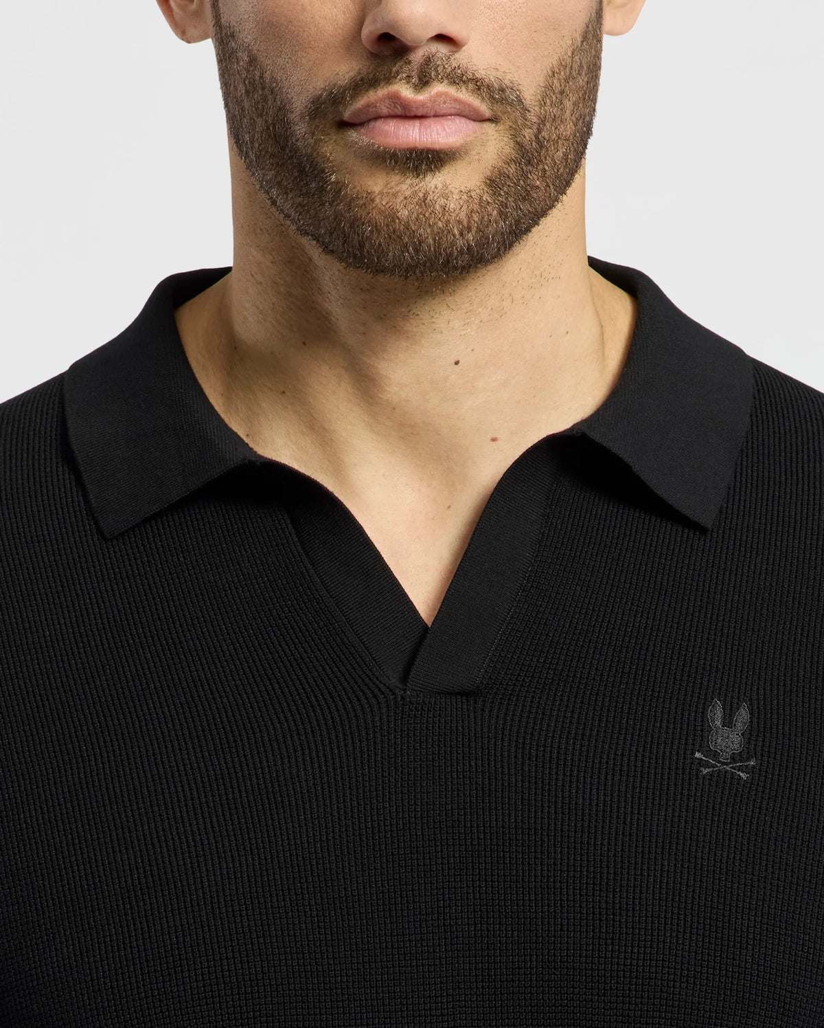 A man wears a black 100% cotton ribbed Psycho Bunny MENS LEETON JOHNNY COLLAR KNIT POLO with an embroidered Bunny emblem on the chest. The breathable short-sleeved shirt is set against a plain white background, showing only his torso and part of his face.
