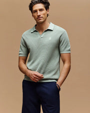 A man with short, wavy dark hair wears the Psycho Bunny MENS LEETON JOHNNY COLLAR KNIT POLO (B6E562F200), a slim fit light green shirt with an embroidered chest Bunny, paired with dark trousers, standing against a beige background.