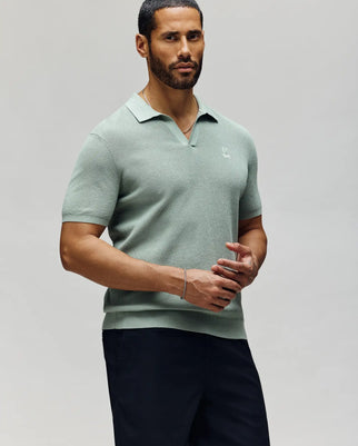 A man with short dark hair and a beard wears the Psycho Bunny MENS LEETON JOHNNY COLLAR KNIT POLO (B6E562F200) in light green and dark pants, standing against a plain light background with a neutral expression and hands together.