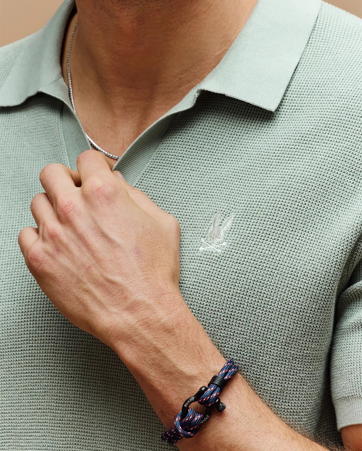 A person wears the Psycho Bunny Mens Leeton Johnny Collar Knit Polo (B6E562F200) in light green, featuring an embroidered chest Bunny, paired with a silver chain necklace and blue-and-black knotted bracelet while holding the necklace.