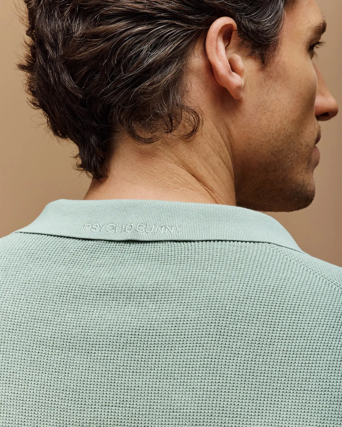 A man is seen from behind in a slim fit light green MENS LEETON JOHNNY COLLAR KNIT POLO by Psycho Bunny, featuring subtle logo embroidery on the collar against a warm, neutral background.