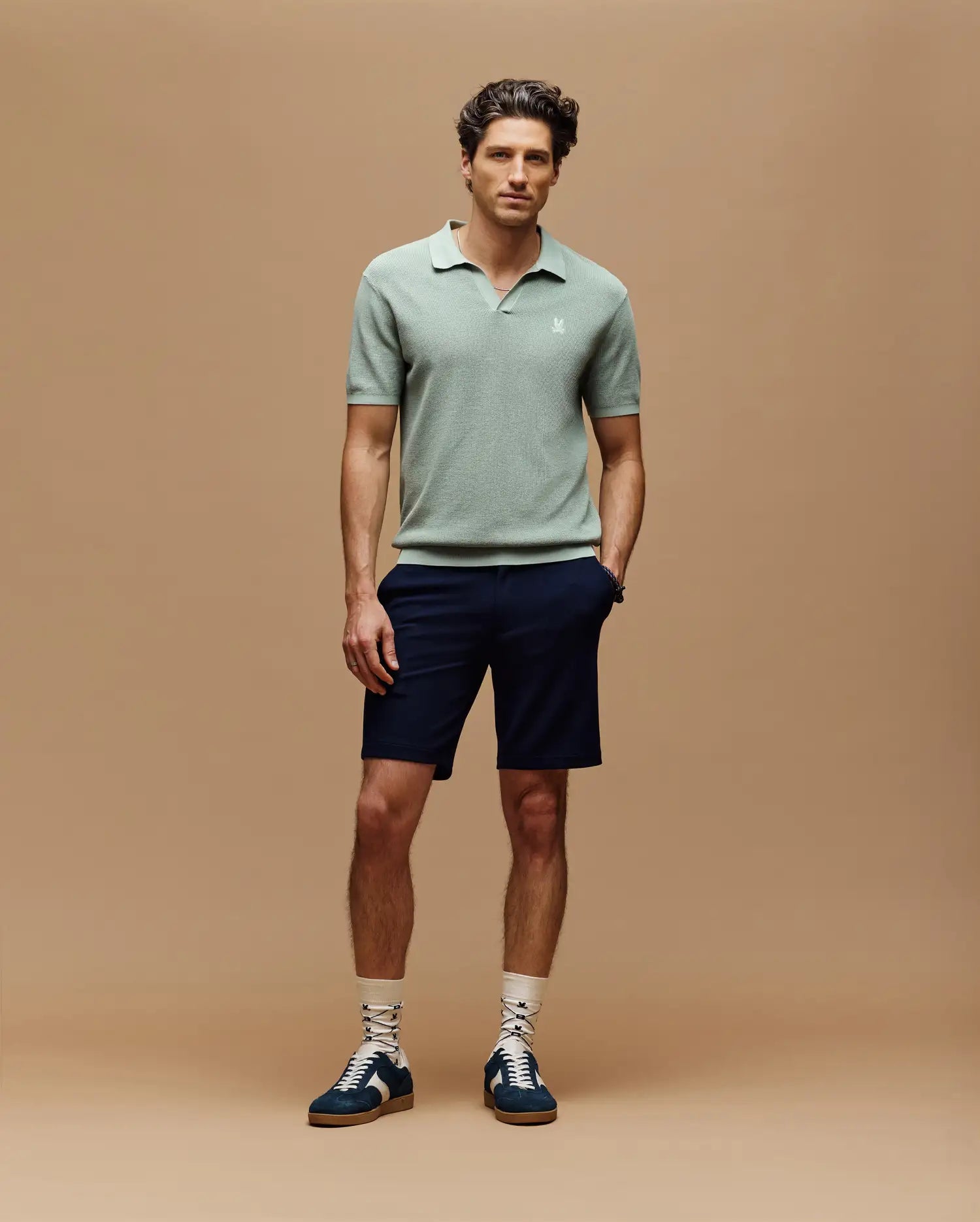 A man with short, styled hair stands against a beige background, wearing the Psycho Bunny MENS LEETON JOHNNY COLLAR KNIT POLO in light green, navy shorts, white patterned socks, and navy sneakers with white laces. His hands are in his pockets.