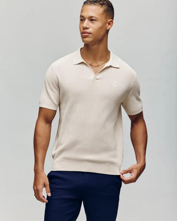 A man stands against a plain background in a Psycho Bunny MENS LEETON JOHNNY COLLAR KNIT POLO - B6E562F200, showcasing a relaxed vibe. The light beige short-sleeve polo pairs with dark blue pants, and he looks to the side exuding ease and comfort in his breathable ensemble.